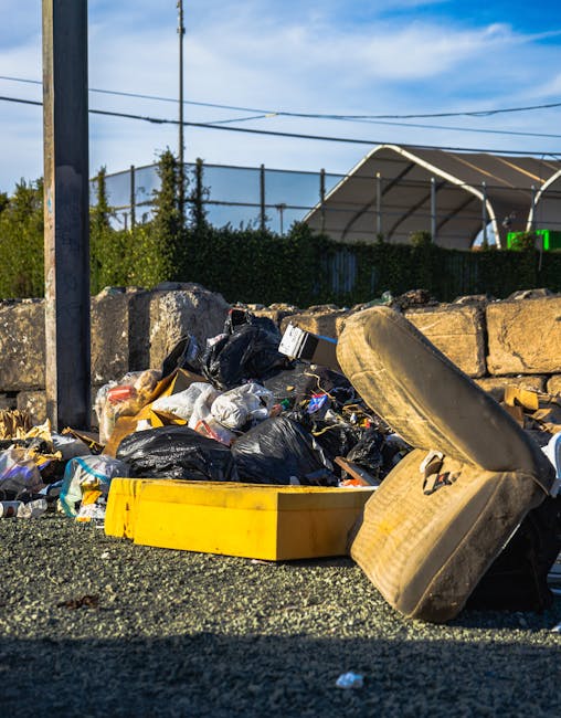 A collection of illegally dumped rubbish on a gravel surface in an outdoor area, featuring black plastic trash bags, a yellow plastic container, and a single old, dirty tan-colored car seat with visible fabric wear and foam padding. The waste is piled up against a low stone wall constructed of irregularly shaped, weathered stone blocks. In the background, there are green shrubs and trees behind a metal fence with vertical bars, and a utility pole with multiple power lines. Overhead, the sky is partly cloudy with patches of blue visible. The scene suggests an informal waste disposal site, highlighting the need for professional rubbish removal services, such as those provided by Waste Clearance Finchley, to handle private or independent waste clearance tasks in the area.