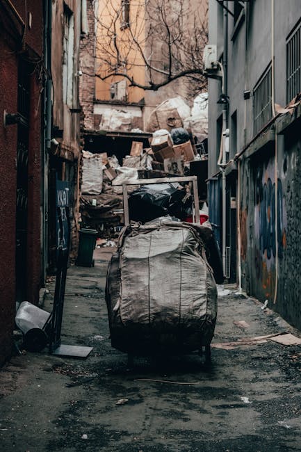 In the foreground of the image, multiple large clear plastic rubbish bags filled with a variety of waste, including textiles, paper, and mixed materials, are piled on a brick pavement. Behind the bags, three workers dressed in black uniforms with brown accents, gloves, and protective headgear are actively engaged in a waste collection task; two are bent down, possibly sorting or lifting debris, while the third stands, reaching towards a stack of boxes or equipment near a storefront. The background features a commercial setting, with a red and white illuminated sign on the left, and a shop display with Japanese-style paper lanterns. Nearby, stainless steel bins with black lids are visible, along with a blue plastic container, indicating waste handling within an urban environment. The scene appears to be part of an independent rubbish collection service, highlighting the process of on-site clearance of bulky waste materials, consistent with private waste management activities conducted by Waste Clearance Finchley.