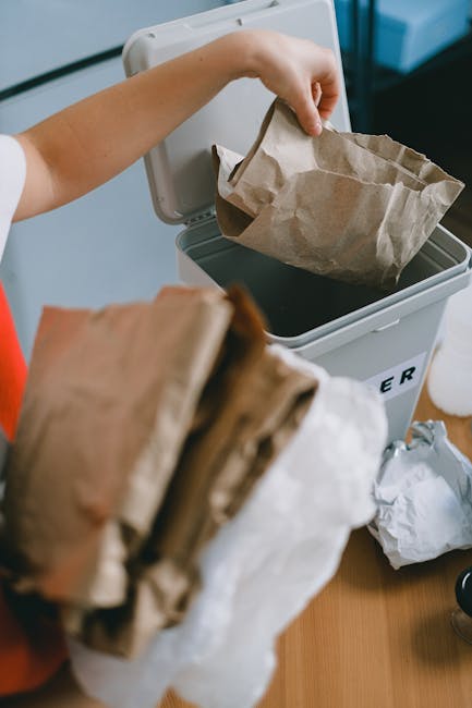 A close-up view of a person's hand placing a crumpled brown paper bag into a white waste bin with a black plastic liner, situated on a wooden surface. In the foreground, there are additional crumpled paper bags held by the individual, with some appearing torn or folded. The background shows additional waste collection materials, including more paper bags and a few miscellaneous items, indicating a waste disposal process possibly related to private rubbish collection near flats or residential buildings. The scene highlights an organized approach to rubbish disposal, consistent with independent collection or on-site clearance services. The waste bin is positioned outdoors or in a semi-covered area, with natural light illuminating the scene and emphasizing the textures of the crumpled paper and the smooth surface of the bin. Waste Clearance Finchley provides these services to facilitate efficient rubbish removal and proper waste handling in urban residential environments, supporting alternative disposal methods outside of local authority collection. The setting appears clean and orderly, emphasizing responsible waste management practices.