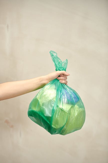 A close-up view of a person's hand placing a crumpled brown paper bag into a white waste bin with a black plastic liner, situated on a wooden surface. In the foreground, there are additional crumpled paper bags held by the individual, with some appearing torn or folded. The background shows additional waste collection materials, including more paper bags and a few miscellaneous items, indicating a waste disposal process possibly related to private rubbish collection near flats or residential buildings. The scene highlights an organized approach to rubbish disposal, consistent with independent collection or on-site clearance services. The waste bin is positioned outdoors or in a semi-covered area, with natural light illuminating the scene and emphasizing the textures of the crumpled paper and the smooth surface of the bin. Waste Clearance Finchley provides these services to facilitate efficient rubbish removal and proper waste handling in urban residential environments, supporting alternative disposal methods outside of local authority collection. The setting appears clean and orderly, emphasizing responsible waste management practices.