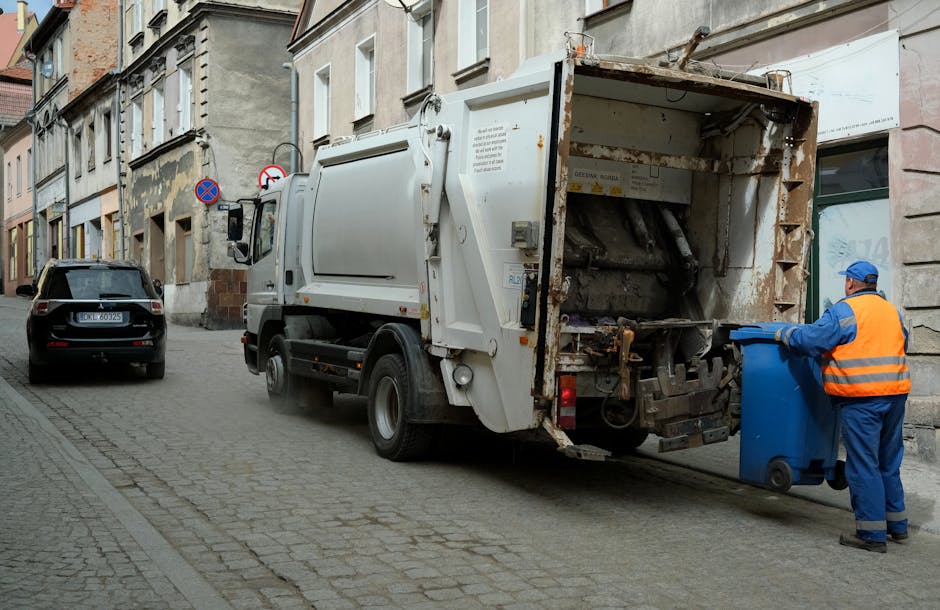 A waste collection truck with a white body and rusted rear section is stationed on a narrow city street beside a row of old, multi-story buildings with stone and brick facades. The truck is positioned slightly to the right of the image, with its rear door open, revealing the interior loading mechanism used for rubbish collection, which appears to have dirt and wear on its surfaces. A worker wearing a blue uniform, orange high-visibility vest, and a blue cap is standing on the right side of the truck, emptying a blue wheeled waste bin into the vehicle. The street has a cobblestone surface with a parked black car visible ahead of the truck and a no-parking sign attached to the building on the left. The scene suggests an active waste collection process, characteristic of regular, independent rubbish removal services, with an emphasis on urban environmental management and private waste handling in the area served by Waste Clearance Finchley.