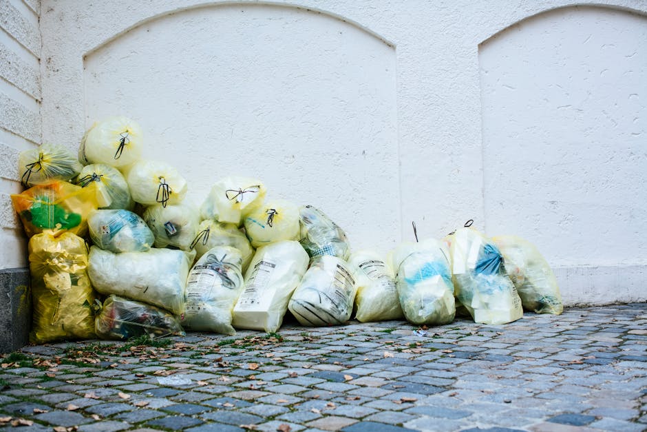 A collection of various rubbish bags piled against a white, textured exterior wall on a cobblestone pavement. The bags are made of transparent and colored plastic, predominantly yellow, white, and blue, and are tied at the top with black or yellow drawstrings. Some bags contain paper, plastic, and other household waste, with visible labels and packaging details. The bags are arranged in a haphazard manner, with some leaning against the wall and others stacked loosely on the uneven cobblestones, which display a mixture of grey, pink, and brown tones with scattered dried leaves and small debris. The scene suggests an outdoor waste collection point, possibly part of private rubbish removal services such as Waste Clearance Finchley, which handles on-site clearance and alternative waste disposal methods, especially in areas where reliance on local authority services is limited. The overall setting is calm and clean, without visible signs of overflowing waste or environmental spillages, reflecting an organized effort to manage household refuse outside residential premises.