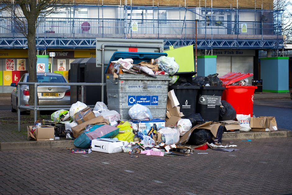 A waste collection truck with a white body and rusted rear section is stationed on a narrow city street beside a row of old, multi-story buildings with stone and brick facades. The truck is positioned slightly to the right of the image, with its rear door open, revealing the interior loading mechanism used for rubbish collection, which appears to have dirt and wear on its surfaces. A worker wearing a blue uniform, orange high-visibility vest, and a blue cap is standing on the right side of the truck, emptying a blue wheeled waste bin into the vehicle. The street has a cobblestone surface with a parked black car visible ahead of the truck and a no-parking sign attached to the building on the left. The scene suggests an active waste collection process, characteristic of regular, independent rubbish removal services, with an emphasis on urban environmental management and private waste handling in the area served by Waste Clearance Finchley.