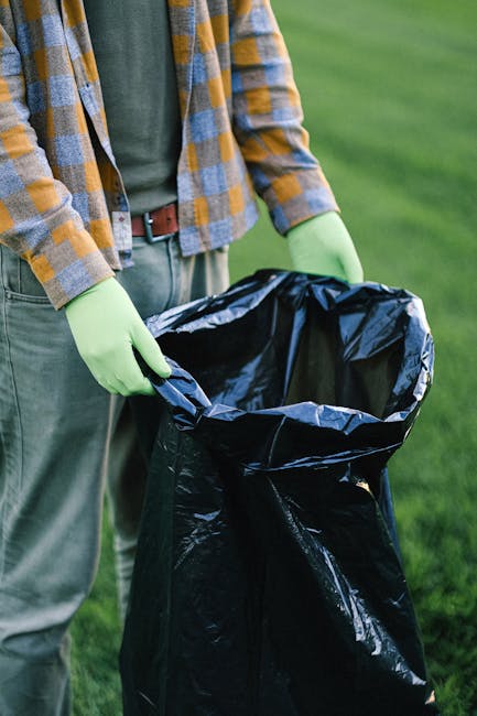 The image shows a person standing outdoors on a grassy area, holding open a large black rubbish bag made of plastic with their left hand. The individual is wearing a green long-sleeve shirt, a plaid jacket in shades of yellow, grey, and brown, and green rubber gloves, indicating a clean-up or waste collection activity. The person's waist is partly visible, showing grey trousers with a brown belt. The background consists of a well-maintained, vibrant green lawn, suggesting a garden, park, or outdoor setting suitable for rubbish removal or environmental care efforts. The scene emphasizes proper waste handling and environmental responsibility, aligning with services offered by waste clearance providers. The lighting is natural, highlighting the textures of the plastic rubbish bag and the fabric of the clothing. This visual depiction is relevant to private waste disposal, rubbish collection, and on-site clearance activities undertaken by Waste Clearance Finchley, supporting efficient rubbish removal in residential or community spaces without shown waste materials.