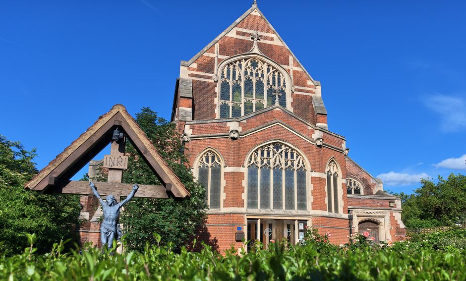 A historic red-brick church with large stained-glass windows and detailed architectural features stands prominently under a bright blue sky. In the foreground, a small wooden structure with a triangular roof is visible, housing a metal crucifix depicting Jesus Christ with arms outstretched and a sign reading 'INRI' above the figure. The church is surrounded by well-maintained greenery, including bushes and trees, which partially obscure the lower portion of the building. The scene is illuminated by natural sunlight, casting clear shadows and highlighting the textures of the brickwork and glass. This setting, combining religious architecture and outdoor surroundings, may contrast with private or on-site waste clearance activities, as part of an independent rubbish removal or maintenance service, such as those offered by Waste Clearance Finchley, to manage debris or outdoor waste in similar environments.