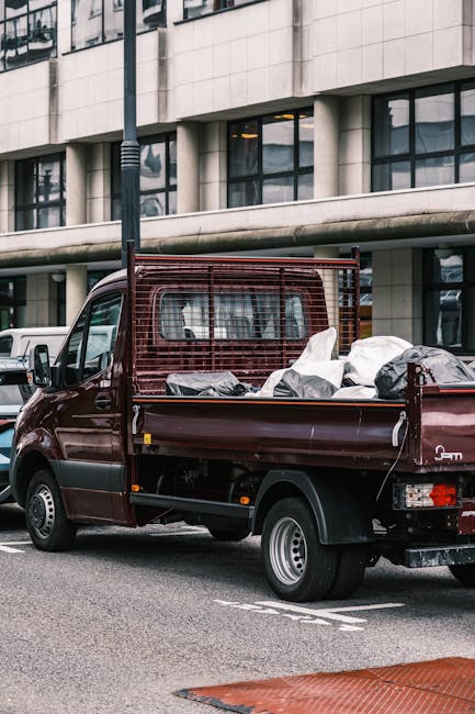 A dark red flatbed pickup truck parked on an urban street in front of a modern building with large glass windows and concrete paneling. The truck's bed is loaded with various types of waste including black and white plastic bags filled with discarded materials, possibly household or construction debris. The bags appear to be made of thick plastic. The truck features a metal safety cage at the front of the bed, painted the same dark red color, which helps secure the load during transport. Surrounding the truck, there are other vehicles and a lamppost, indicating a busy city area. The scene is captured in daylight with natural lighting, emphasizing the neutral and professional tone suitable for waste management and rubbish removal services. Waste Clearance Finchley may utilise such vehicles for alternative waste handling or private disposal tasks, highlighting their capacity for on-site clearance in urban environments. The street surface is asphalt with painted markings, and part of a textured, red-colored curb or ramp is visible in the foreground, contributing to the sense of an organized urban setting dedicated to Efficient rubbish collection and waste clearance activities.