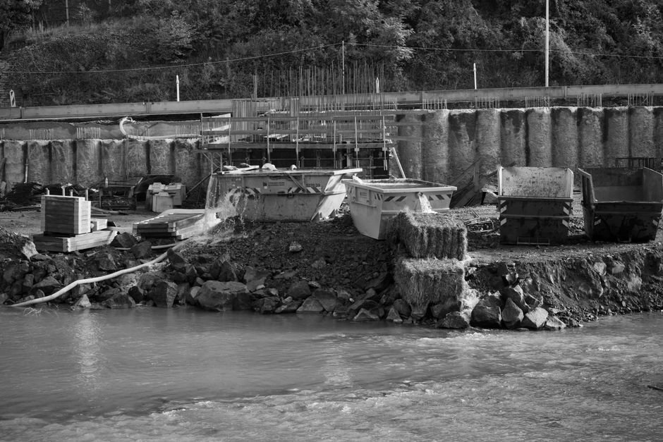 The image depicts a construction or renovation site situated alongside a body of water, with several boats docked on a rocky shoreline in the foreground. The boats are made of fiberglass or plastic, with one featuring a visible cabin and others designed for leisure, all appearing to be in a state of disuse or awaiting maintenance. Adjacent to the boats, there are stacks of wooden planks and construction materials, including concrete blocks and metal rebar, which are positioned on the uneven, soil-covered terrain. To the right, a large metal skip or dumpster is filled with debris, with its open top revealing discarded waste, possibly construction rubbish. Behind the boats and equipment, there is a tall, reinforced concrete retaining wall with steel reinforcement bars protruding from the top, supporting an upper level with a railway track and a guardrail, suggesting proximity to a roadway. The overall scene is monochrome, with natural lighting casting shadows that emphasize the textures of the gravel, rocks, and construction materials, providing a clear visual context relevant to local waste handling and alternative waste disposal methods often used in private site clearance or independent rubbish removal services offered by companies like Waste Clearance Finchley.