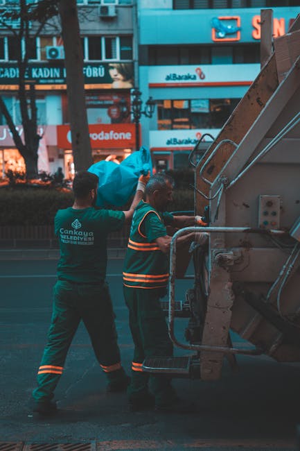 Two waste collection workers dressed in dark green uniforms with reflective orange stripes are standing beside a large waste disposal vehicle on a city street. One worker is holding a large, bright blue tarp or bag, which appears to contain waste materials, while the other is operating the vehicle's controls or loading mechanism. The vehicle is a large, industrial-grade waste truck with visible metal components, hydraulic arms, and a rear compactor section. The background features a cityscape with illuminated commercial signage, including store signs for Vodafone and other businesses, and a mix of modern buildings with glass windows. The scene is lit by ambient street lighting, casting a soft glow on the workers and the waste vehicle, indicating early evening or night-time. The environment shows a typical urban setting where private waste collection services, such as Waste Clearance Finchley, may conduct on-site clearance or alternative rubbish collection outside of local authority collection schedules.